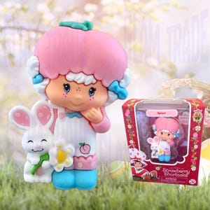 May include: A Strawberry Shortcake Apricot figurine featuring a character with pink hair, a white apron, and blue shoes, standing next to a white rabbit holding a yellow flower. The figurine is also shown inside its original red and white packaging.
