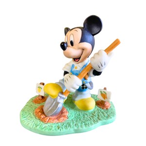 Peut inclure: Une figurine en c&eacute;ramique de Mickey Mouse portant une salopette, tenant une pelle et creusant dans un jardin. La figurine est pos&eacute;e sur une base verte avec de la terre brune et des fleurs jaunes.
