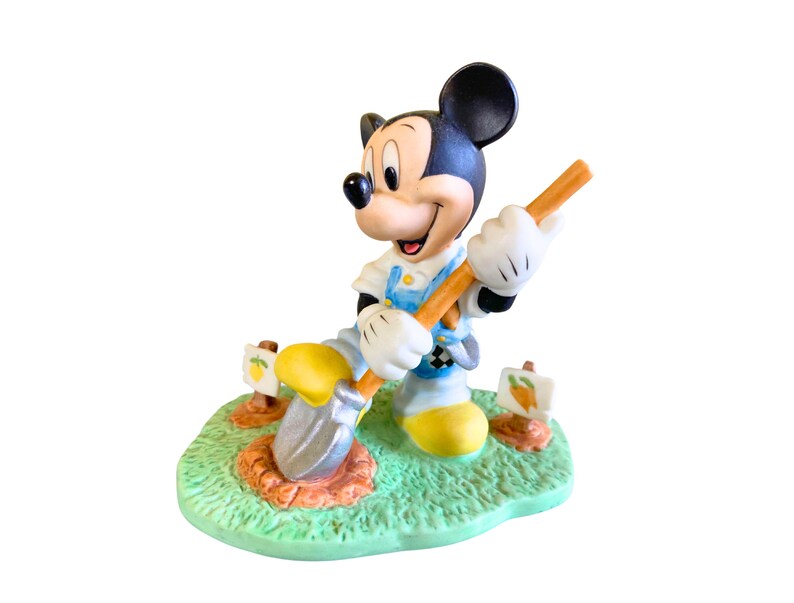 Peut inclure: Une figurine en c&eacute;ramique de Mickey Mouse portant une salopette, tenant une pelle et creusant dans un jardin. La figurine est pos&eacute;e sur une base verte avec de la terre brune et des fleurs jaunes.