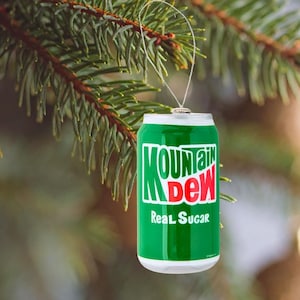 May include: A green and white Mountain Dew can ornament with the words "MOUNTAIN DEW" in green and red, and "Real Sugar" in white. The ornament hangs from a silver string on a Christmas tree branch.