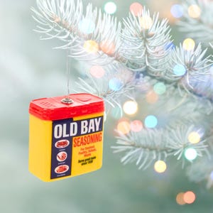 May include: A yellow and red Old Bay Seasoning box ornament hanging from a Christmas tree branch. The ornament features the brand's logo and text, with illustrations of seafood and meat. The text reads "For Seafood, Poultry, Salads, Meats. Same great taste since 1939."