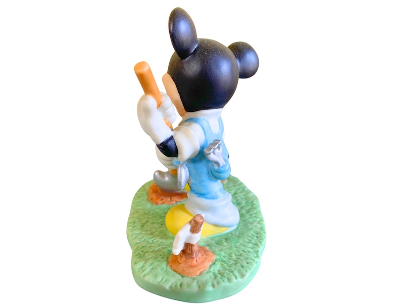 Peut inclure: Une figurine en c&eacute;ramique de Mickey Mouse habill&eacute; d'une salopette bleue et d'une chemise blanche, tenant un club de golf. Il est debout sur une base verte avec une balle de golf et un club de golf.