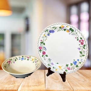 May include: A white ceramic plate and bowl set with a floral design. The plate is displayed on a wooden stand. The floral pattern includes pink, blue, and yellow flowers with green leaves. The bowl has a dark gray base.
