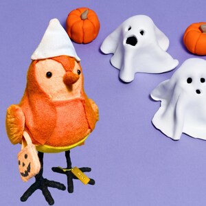 May include: A Halloween-themed display featuring an orange bird wearing a white hat and holding a pumpkin-shaped bag. Two white ghost figures and two orange pumpkins are also present, all set against a purple background.