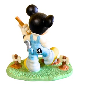 Peut inclure: Une figurine en c&eacute;ramique de Mickey Mouse portant une salopette bleue et une chemise blanche, tenant une pelle et debout sur une base verte avec des piquets en bois brun.