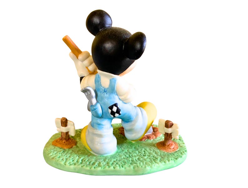 Peut inclure: Une figurine en c&eacute;ramique de Mickey Mouse portant une salopette bleue et une chemise blanche, tenant une pelle et debout sur une base verte avec des piquets en bois brun.