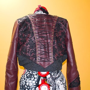 Op de afbeelding: Een kort, bordeauxrood leren jack met zwarte kantdetails en een gespeld rugpaneel. Het jack is op een mannequin met een zwart-wit gepatroneerd torso.