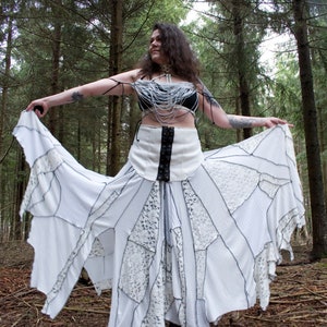 May include: A woman wearing a white skirt with black lace detailing and a white beaded top. She is standing in a woodland setting with trees in the background.