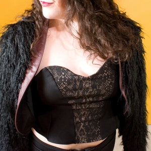 May include: A black bustier top with a lace and snake-skin pattern, worn with a black faux fur jacket with a burgundy lining. The outfit is set against a yellow background.