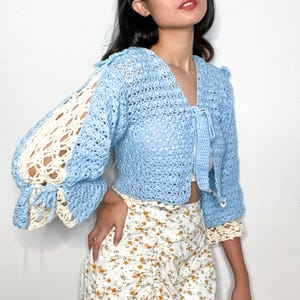 Peut inclure: Un cardigan en crochet bleu clair avec une bordure en crochet blanc. Le cardigan a un nœud à l'avant et est court. Les manches sont longues et ont une forme de cloche.