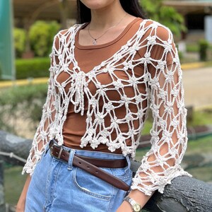 Könnte beinhalten: Weiße Häkeljacke mit zartem Blumenmuster. Die Jacke hat eine verkürzte Länge und lange Ärmel. Sie wird über einem braunen Tanktop und einer blauen Jeans getragen.