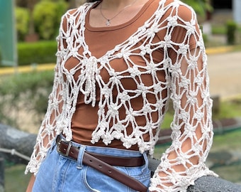 May include: White crochet cardigan with a delicate floral pattern. The cardigan has a cropped length and long sleeves. It is worn over a brown tank top and blue jeans.