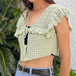 May include: A light green crochet top with a large, ruffled collar and short sleeves. The top is tied at the front with a bow.