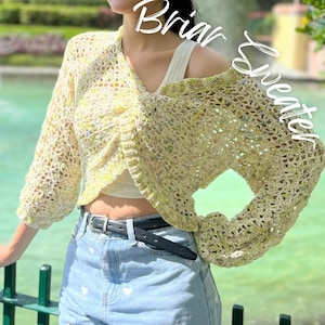 May include: A crocheted cream and yellow sweater with a loose fit and long sleeves. The sweater has a V-neck and a cropped length. The sweater is worn over a white tank top and paired with blue denim shorts.