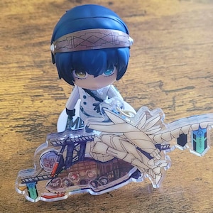 May include: A character figurine with blue hair and a brown headband, dressed in white, sits above a clear acrylic charm. The charm depicts a detailed illustration of a vehicle with wings and the text "Genshin Impact".