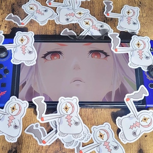 May include: A blue Nintendo Switch controller with a screen displaying a female anime character with red eyes. White stickers of a cartoon bear with a star and a cigarette are scattered around the console on a wooden surface.