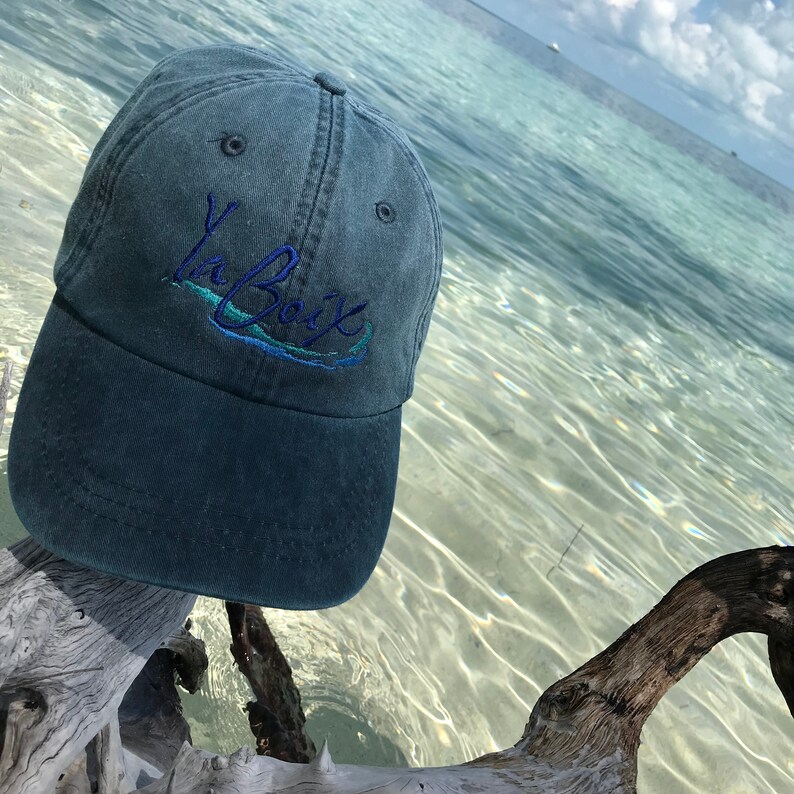 K&ouml;nnte beinhalten: Dunkelblaue Baseballkappe mit dem gestickten Text "La Baix" in Blau und T&uuml;rkis. Die Kappe liegt auf einem St&uuml;ck Treibholz, mit einem klaren Ozean und Himmel im Hintergrund.