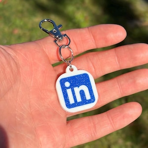 May include: A white and blue LinkedIn logo keyring with a silver clasp. The keyring features the LinkedIn logo in blue against a white background. The keyring is attached to a silver metal clasp.