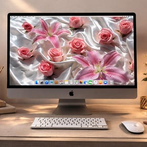 May include: A modern desktop computer with a white keyboard and mouse sits on a desk. The computer screen displays a floral wallpaper with pink roses and lilies. A lit candle, a desk lamp, and a vase of roses are also on the desk.