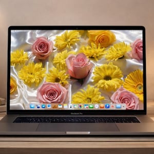 May include: A silver laptop displays a floral wallpaper of pink roses and yellow chrysanthemums. The laptop is on a wooden desk with a candle, a vase of roses, and a desk lamp. The scene has a warm, inviting ambiance.