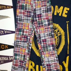 May include: Multi-colored patchwork pants with a plaid pattern in red, yellow, green, and blue. The pants are hanging on a wooden hanger. The background includes pennants and a navy blue banner with yellow text.