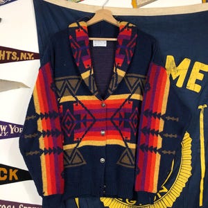 May include: A navy blue cardigan with a shawl collar and a colourful geometric pattern. The pattern features red, orange, yellow, brown, and purple stripes and diamonds. The cardigan has a button closure.