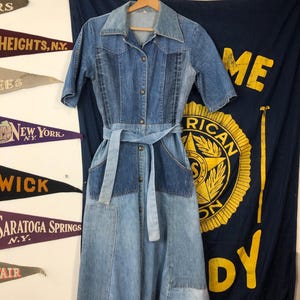Puede incluir: Un vestido de mezclilla azul con un diseño de patchwork. El vestido tiene cuello, mangas cortas y cierre de botones en la parte delantera. Cuenta con un cinturón y dos bolsillos de parche.