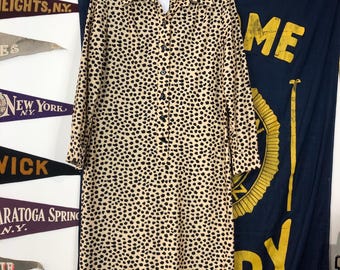 Puede incluir: Una camisa de botones de manga larga de color beige con un estampado de lunares negros y blancos. La camisa tiene cuello y cierre de botones en la parte delantera. La camisa está colgada de una percha.