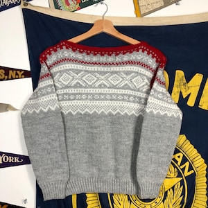 Puede incluir: Un suéter gris y blanco con un patrón y un borde rojo alrededor del cuello. El suéter está hecho de una tela suave y tejida.