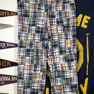May include: Patchwork trousers with a plaid design in blue, green, red, and yellow. The trousers are displayed on a hanger. The background features pennants and a navy blue banner.