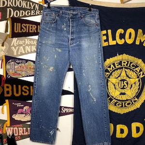 Puede incluir: Vaqueros de mezclilla azul descolorido con salpicaduras de pintura, colgados de una percha de metal. Los vaqueros tienen bragueta con botones y diseño clásico de cinco bolsillos. El fondo incluye banderines vintage y una pancarta con el texto "WELCOME".