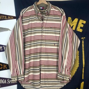 Può includere: Una camicia a maniche lunghe con bottoni e strisce orizzontali beige, rosse e nere. La camicia ha un colletto classico e una singola tasca sul petto.