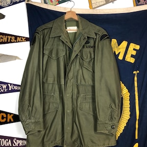 May include: Olive green U.S. Army jacket with multiple pockets and the words "S. ARMY" on the chest. The jacket is hanging on a wooden hanger. Patches are visible on the sleeves. The background includes pennants and a blue banner.
