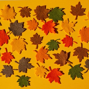 May include: A collection of felt autumn leaves in various shades of red, orange, yellow, brown, and green. The leaves are arranged on a yellow background.