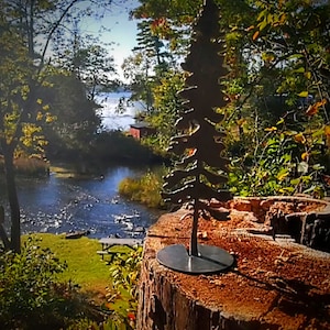 May include: A metal silhouette of a pine tree on a tree stump. The tree is dark gray and the stump is brown. The background is a blurry image of a river and trees.
