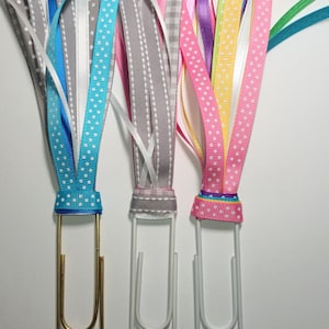 May include: Three gold, silver, and white paper clips with colorful ribbon bows attached. The ribbons are pink, gray, and blue with white polka dots.