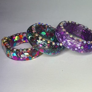 May include: Three colorful resin rings. One ring features a pattern of colorful circles, another is filled with green and pink glitter, and the third is purple with glitter. Each ring is adorned with small, clear crystals.