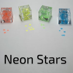 May include: Six small clear cubes filled with neon star-shaped glitter. The cubes are arranged in a semi-circle on a white surface. The glitter colors are pink, orange, yellow, green, blue, and purple. The text "Neon Stars" is written below the cubes.
