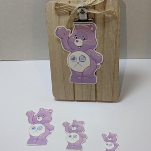 May include: A wooden clipboard with a metal clip and a jute bow, featuring a purple Care Bear sticker. Three additional Care Bear stickers of varying sizes are also present, all with the same design.