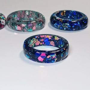 May include: Five colorful resin rings with embedded glitter. The rings are in shades of blue, green, pink, and white.