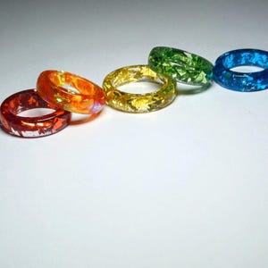 May include: Five colorful resin rings with glitter embedded in the resin. The rings are in shades of red, orange, yellow, green, and blue.