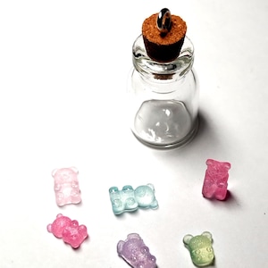 May include: A small, clear glass bottle with a cork stopper and a metal ring on top. Scattered around the bottle are several colorful gummy bear candies in shades of pink, blue, purple, and green. The candies have a sparkly, translucent appearance.
