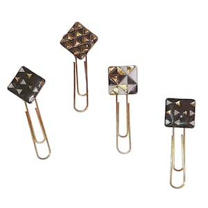 May include: Four gold paper clips with decorative black and white geometric designs. The designs feature gold accents.