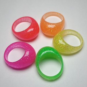May include: Five neon-colored rings with glitter embedded in the resin. The rings are pink, orange, yellow, green, and lime green.