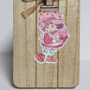 May include: A wooden clipboard with a metal clip and a jute bow. A glittery sticker of a cartoon girl in a pink dress and hat is attached to the clipboard. The girl is holding a pink cat.
