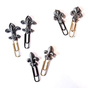 May include: A collection of decorative paper clips featuring a fleur-de-lis design. The clips have a metallic gray finish and are attached to either gold or black paper clips. The items are arranged on a white surface.
