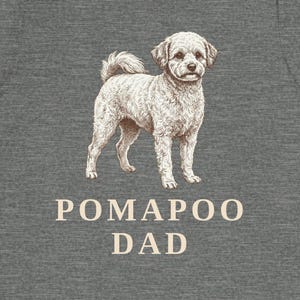 May include: A heather gray short-sleeve t-shirt featuring a detailed illustration of a light-colored Pomapoo dog standing in the center. Below the dog, the text POMAPOO DAD is printed in a clean, light-colored sans-serif font on the chest area.