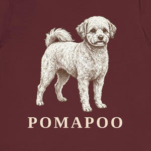 May include: A maroon short-sleeved t-shirt featuring a detailed white line-art illustration of a standing Pomapoo dog centered on the chest. Below the dog illustration, the word POMAPOO is printed in a clean, light-colored sans-serif font.