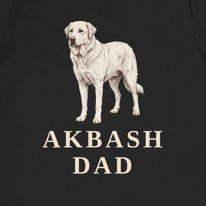 May include: Black t-shirt featuring a detailed illustration of an Akbash dog. The dog is white with a light brown outline. Below the dog is the text "AKBASH DAD" in a serif font.