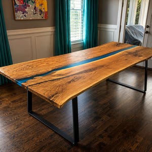 May include: A large, rectangular wooden table with a live edge design. The tabletop is a warm brown with a blue resin river running down the center. The table is supported by black metal legs.